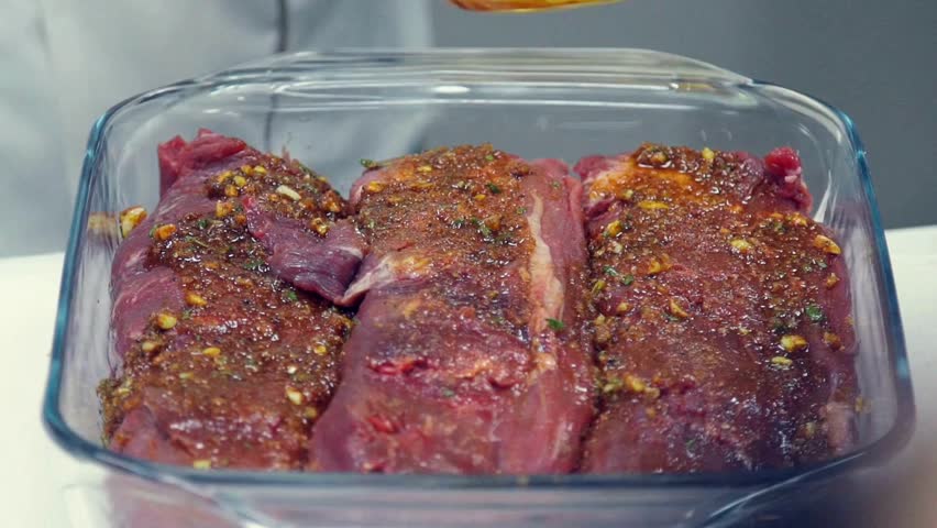 Close up of a cook spreading an herb marinade over raw beef tenderloin in a glass tray, professional culinary preparation for roasting