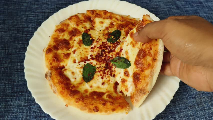 Hand pick and holding Italian margherita pizza slice with simple mozzarella cheese and basil leaves topping from white plate for eating. Delicious pizza served on restaurant table closeup top view.