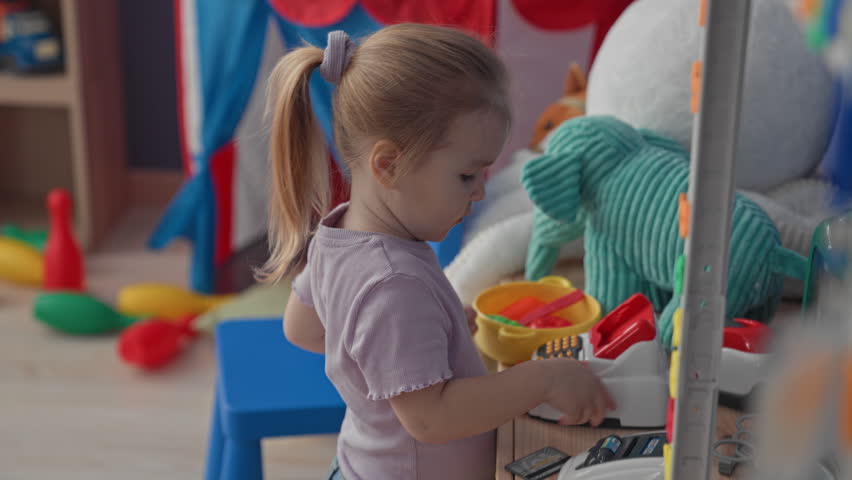 Girl adjusts round toy glasses while pressing buttons on a toy cash register in a playroom building; concentration imagination.