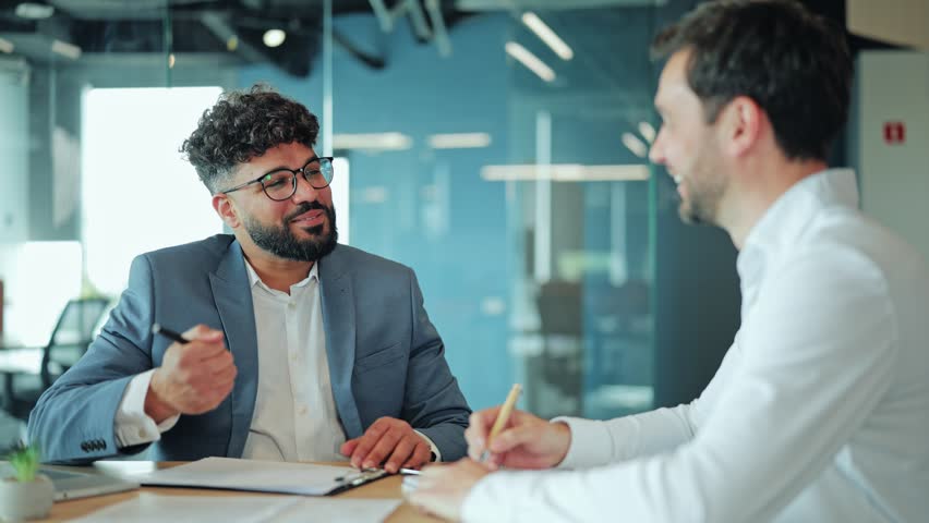 Two diverse businessmen sitting at desk during legal agreement. Middle Eastern man in suit smiling and looking at European colleague or lawyer who is writing on paper or contract document.