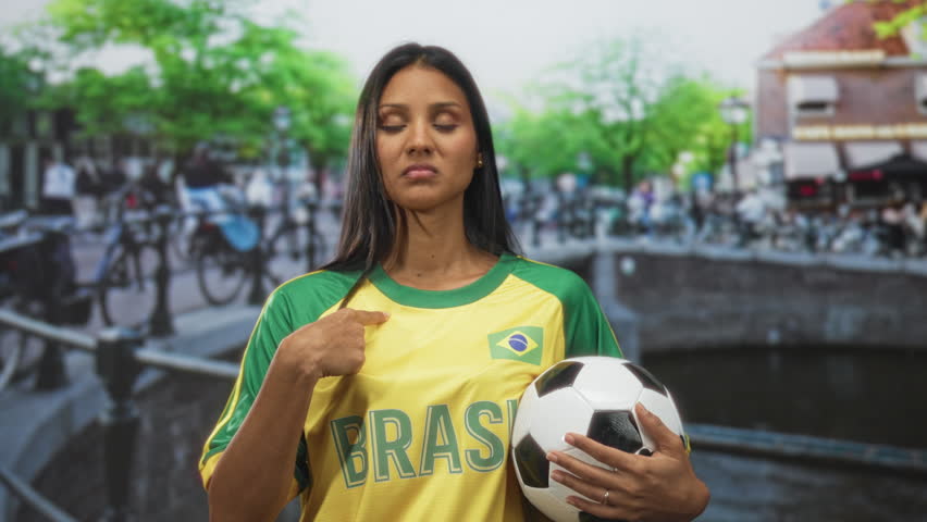 Woman young brunette in brazil t shirt points finger to chest while holding soccer ball on a canal street bridge with bicycles; pride team support.