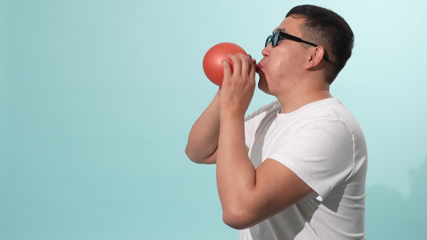Young asian man in sunglasses blowing up a red balloon in studio