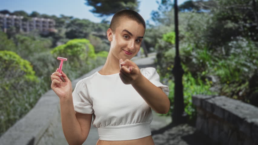 Woman with shaved head holding pink razor and points finger to temple on a park pathway; confidence empowerment.