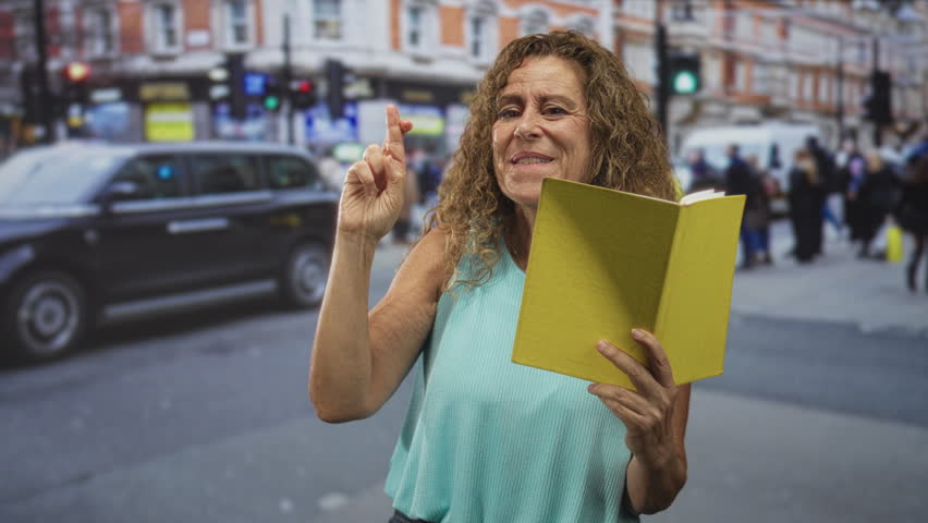 Woman holding yellow book, fingers crossed raised, reading aloud while gesturing on a crowded city street crosswalk amid blurred pedestrians and traffic; hopeful recitation.