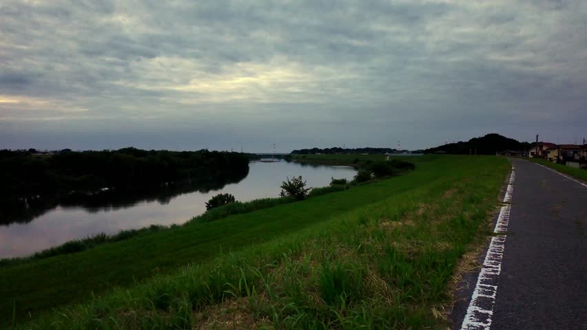 Serene river landscape with a grassy embankment and a paved road under a cloudy sky at dusk