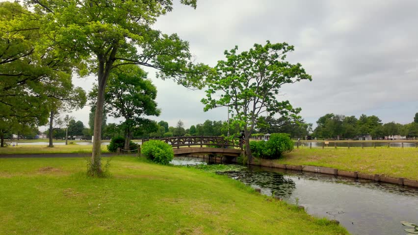Serene park landscape with a wooden bridge over a stream and a person riding a bicycle in the background under a cloudy sky