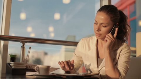beautiful young woman sitting at a table in a cafe, she works with the tablet, talking on the phone and making notes in a notebook. Then stretches his neck and drinks coffee. she was tired