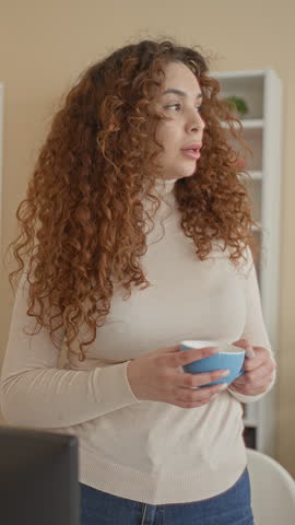 Woman with curly hair holding blue coffee cup near laptop, hands visible, standing beside shelving in studio; calm contemplation.