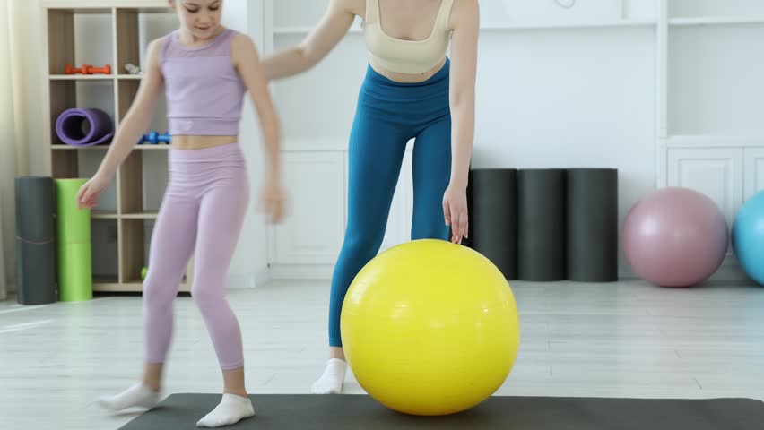 Trainer helping little girl to do exercise with fitness ball indoors, closeup