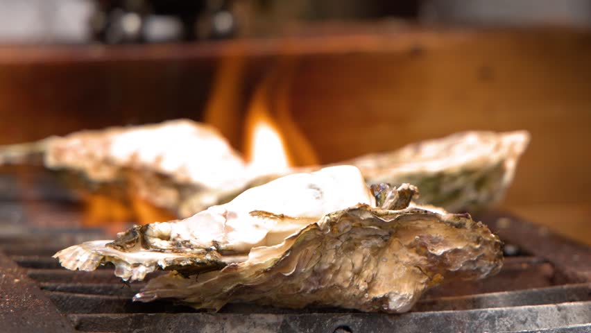 Close-up shot of large oysters cooking on a grill with flickering orange flames in Tokyo.