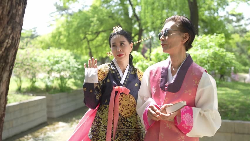 Two women in traditional Korean attire walk through a park. One points at a tree while the other observes. They appear thoughtful and engaged. Ideal for cultural, historical, or travel-themed projects