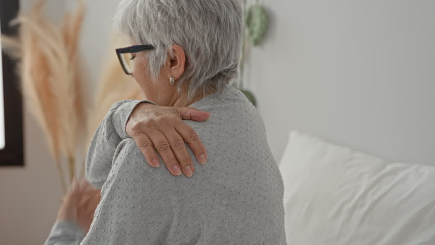 Woman with short grey hair holds her shoulder with hand on a bed in a building, wearing glasses and a long sleeve sweater; pain recovery.