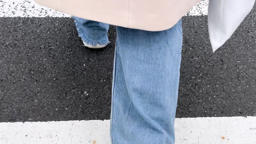 A low angle tracking shot follows a person in blue denim jeans crossing a street