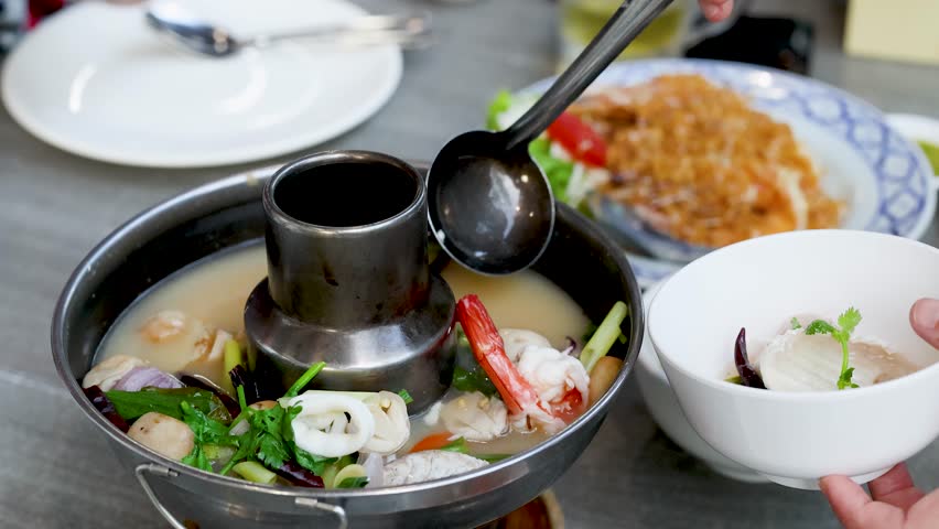 A person uses a metal ladle to serve spicy seafood soup into a white bowl