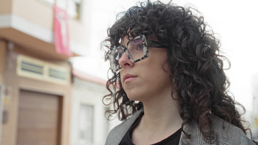 Woman wearing tortoiseshell sunglasses looks sideways on an urban street in front of a building garage door; quiet contemplation.