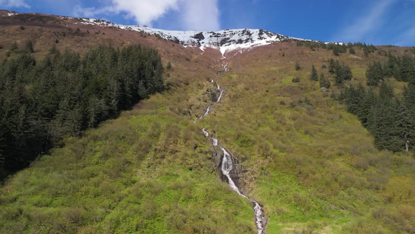A glacial waterfall cascades down a steep mountainside slope near Juneau, Alaska, feeding icy cold water into the landscape below. Surrounded by rugged terrain and lush greenery, it showcases the raw, untouched beauty of Southeast Alaska’s glacial wilderness.