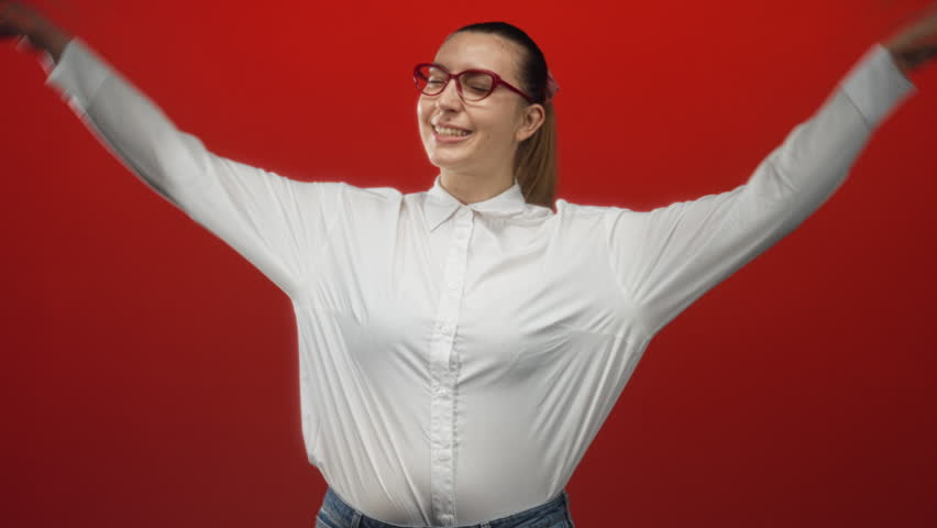 Woman wearing white shirt and red glasses with hands making bunny ears over head in studio; playfulness lighthearted charm.