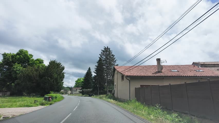 A vehicle travels along a paved road past stone houses and trees under cloudy skies