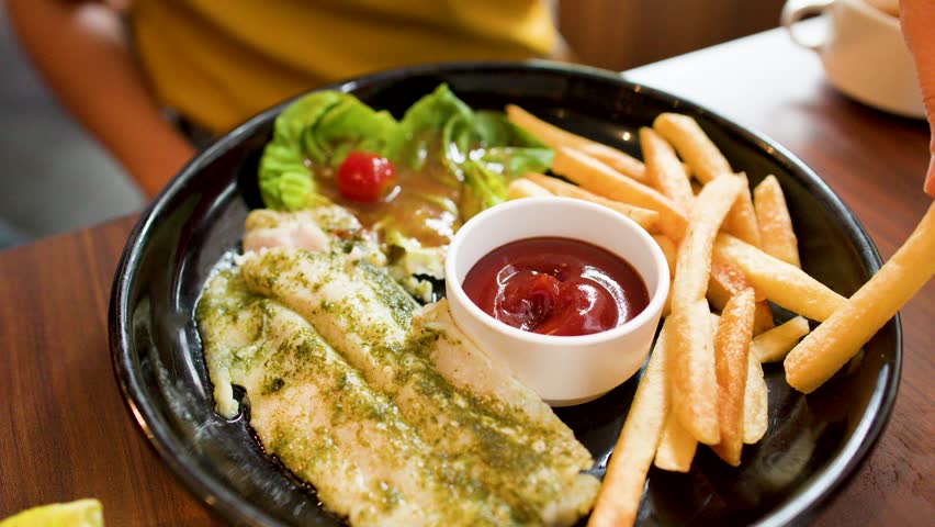 A hand dips a golden french fry into red ketchup beside grilled pesto fish