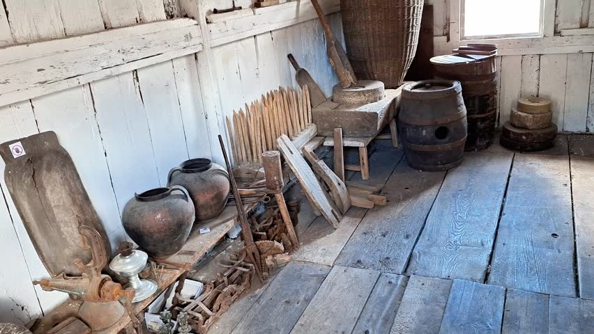 Rustic wooden interior filled with antique household tools, clay pots, barrels, and vintage farm equipment inside a historic rural cabin with natural daylight
