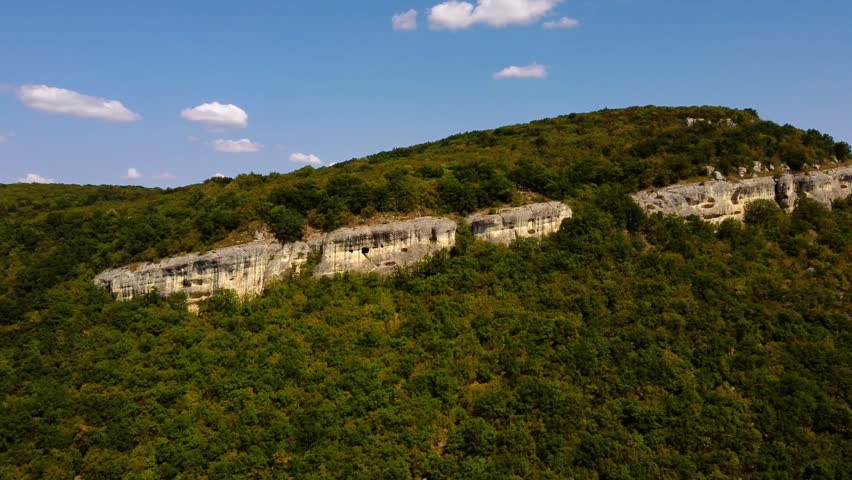 Aerial slow motion tracking shot of limestone cliff and forested hillside under clear blue sky.