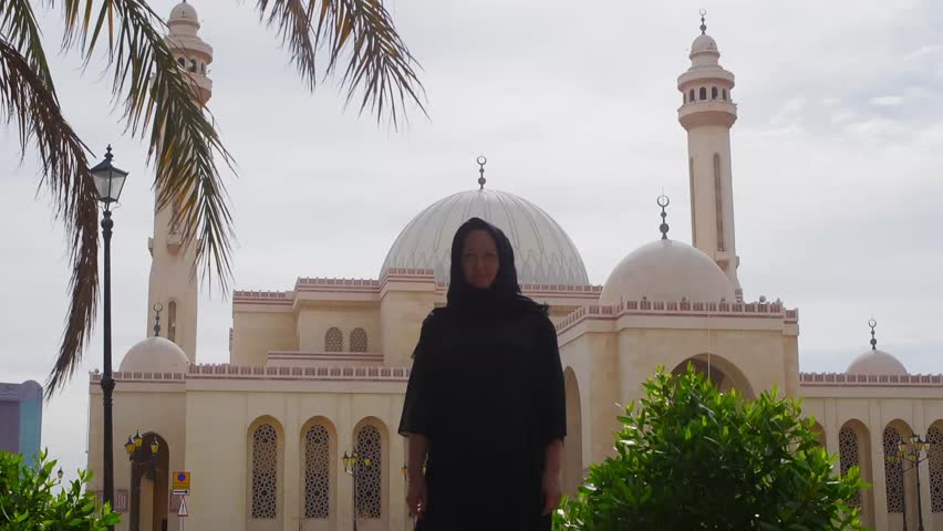 Woman in black abaya poses before ornate mosque featuring white domes, tall minarets, Islamic architecture in tropical setting with palm fronds.