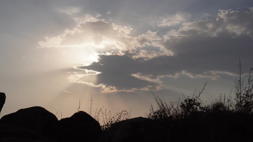 Sun rays breaking through dramatic clouds above a rocky lakeside landscape during golden hour.