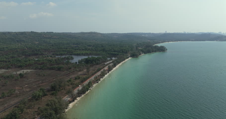 Forward aerial view of a train traveling on tracks beside a coastal road, separated by trees from the turquoise sea along a scenic shoreline.
