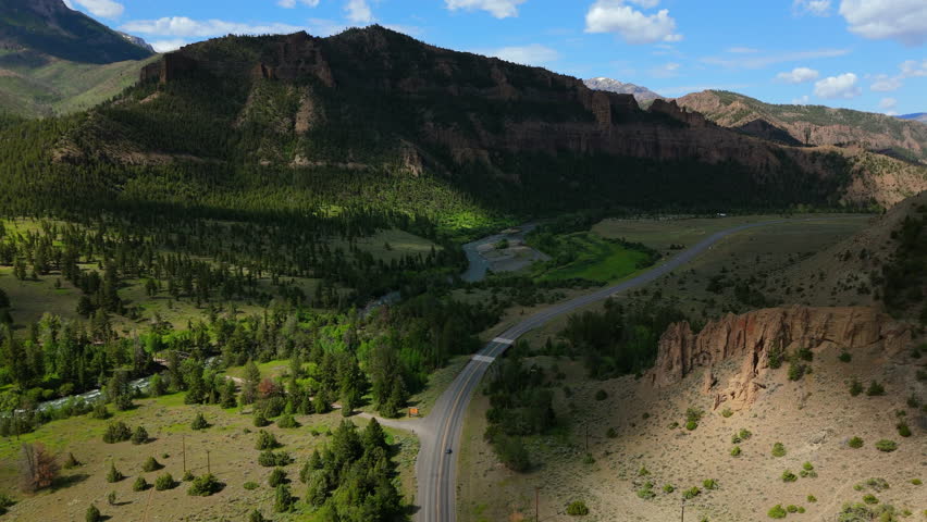 Aerial Montana USA reveals breathtaking mountain valley with dramatic cliffs, winding two-lane highway, lush green meadows dotted with pine trees, and a river flowing beneath bright blue skies.