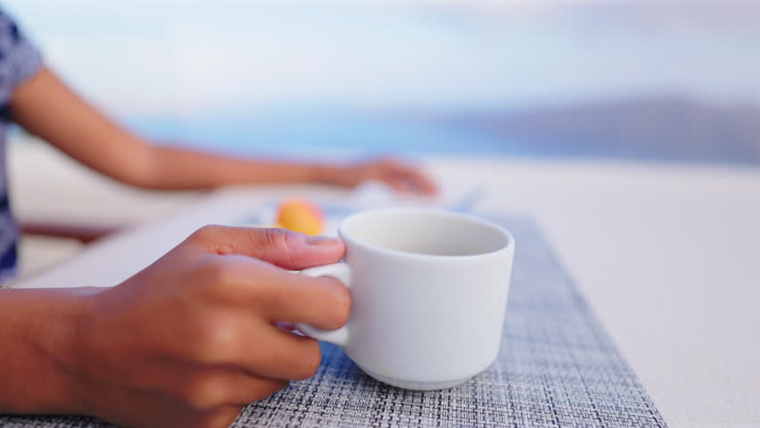 Woman wearing blue sunglasses and embroidered dress sips coffee from white cup at outdoor terrace in Santorini Greece. Blurred Mediterranean Sea and caldera create serene vacation atmosphere.