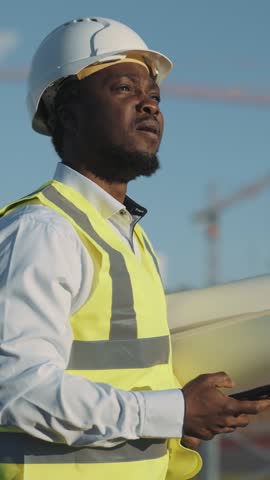 Innovations in architecture and building, vertical portrait of black foreman in modern construction site. Architect controlling work of building cranes, foreman walking in future new districts in town