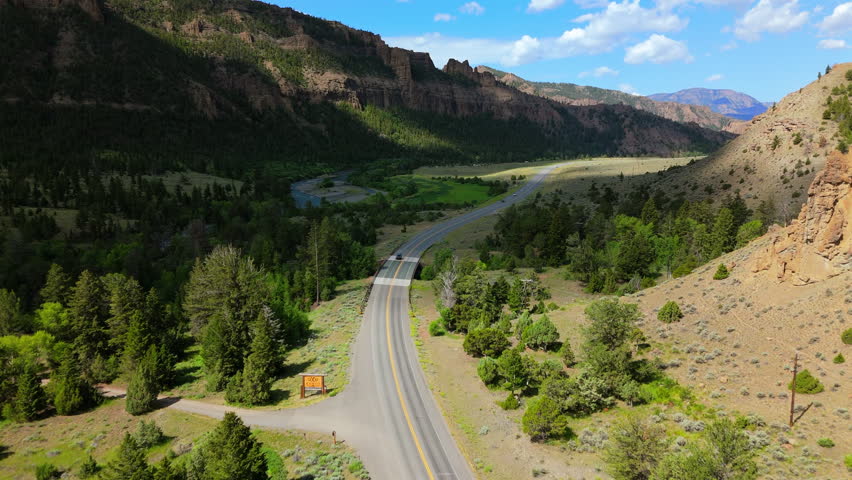 Aerial Montana USA view captures scenic mountain highway winding through lush green valley. Dramatic cliffs and pine forests frame the road as vehicles travel along the river corridor.