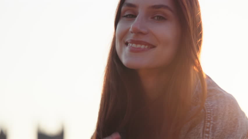 Beautiful young redhead woman with flowing auburn hair smiles radiantly while flashing peace sign and playfully sticking out tongue during warm golden hour sunset with soft backlit glow.