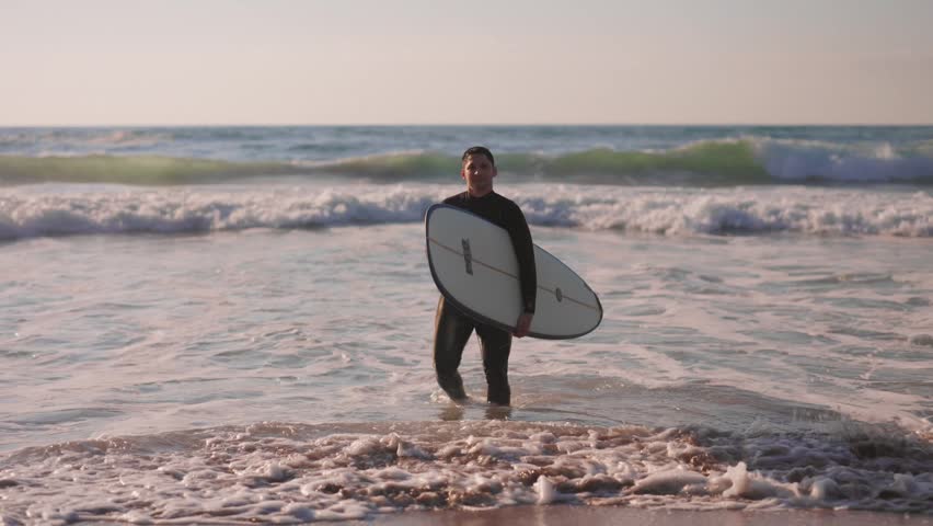 Handsome male surfer in a wetsuit carrying his surfboard and walking out of the ocean water onto the sandy beach after a surfing session during a beautiful and warm summer sunset