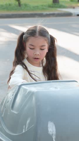 A young Thai girl puts a pink plastic bag into a grey recycling bin outdoors