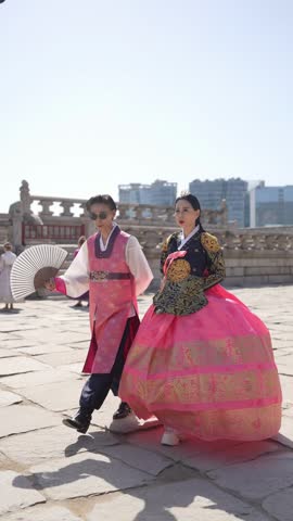 Bright sun shines on ancient stone walkway. Two people in vibrant traditional attire stroll together. Man holds fan, woman carries bouquet, both smile gently. Background shows distant cityscape