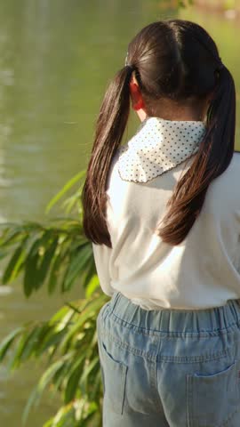 A young girl stands by a lake, tossing food to fish during golden hour sunlight