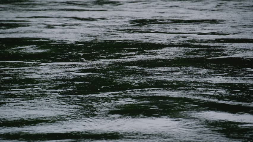 Dark water ripples slowly under gray sky. Surface reflects muted light, creating soft texture. Calm, serene mood evokes quiet nature or melancholic atmosphere. Perfect for films, intros