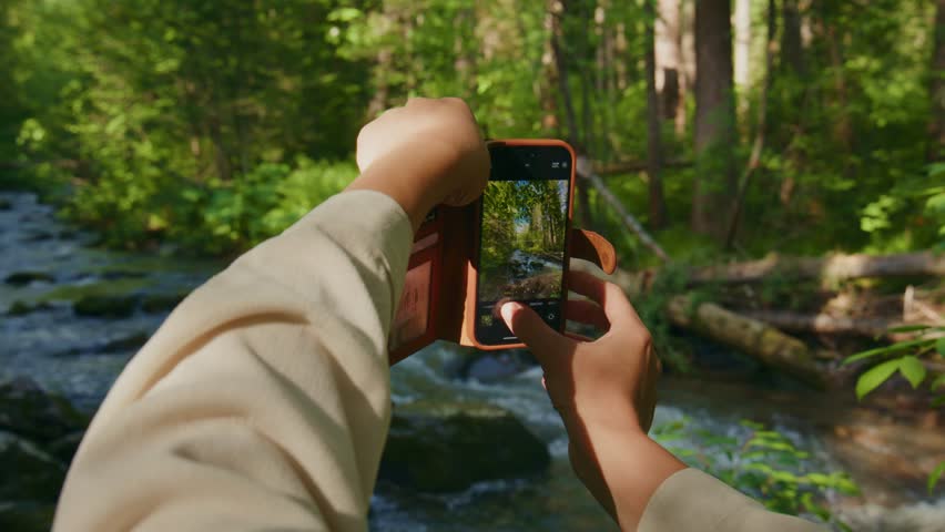 Person captures forest stream with smartphone. Hands steady, focused on framing nature. Sunlight filters through green canopy. Ideal for travel, adventure, or eco-themed content