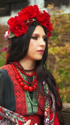 portrait happy girl sitting near house well yard. Young woman slavic beauty ukrainian black dress wreath red beads, pretty face long dark hair. Lady wraps herself in flower warm scarf cold autumn wind