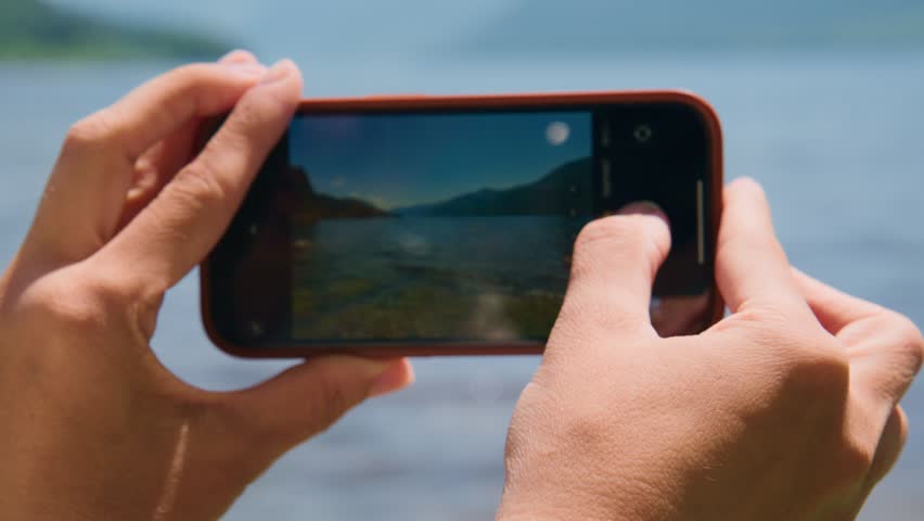 Hands hold smartphone, capturing scenic lake view. Fingers adjust camera focus on screen. Shot shows calm water, distant mountains. Ideal for travel, nature, or tech themes