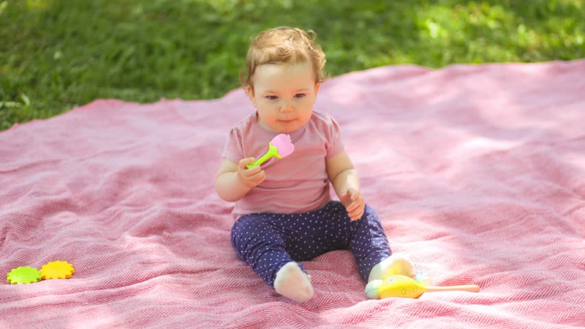 A cute caucasian infant toddler sits on a pink blanket on the green grass, holding and exploring colorful spoons and toys during a bright summer day picnic in a park.
