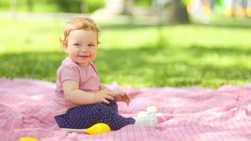 A cheerful caucasian infant toddler in a pink shirt sits on a picnic blanket on the grass, smiling warmly towards the camera with green trees in the blurred background.