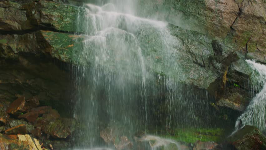 Close-up of a small waterfall flowing over rocky cliff with green moss and wet stone textures. Fresh natural landscape, peaceful outdoor scene with moving water.