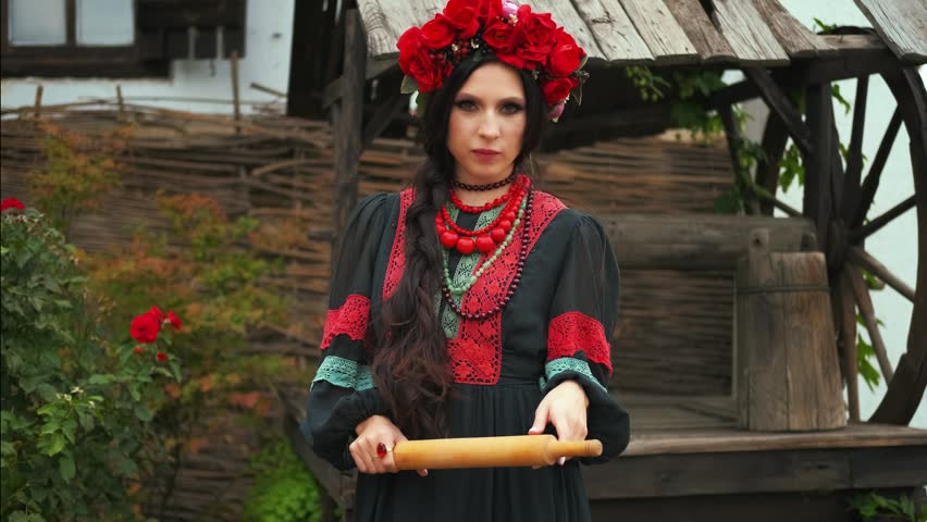 Fantasy portrait warlike evil woman with bat, holding in hands threatening looks at camera. black dress red beads wreath roses crown poppy flowers. angry wife threatens her husband with rolling pin.