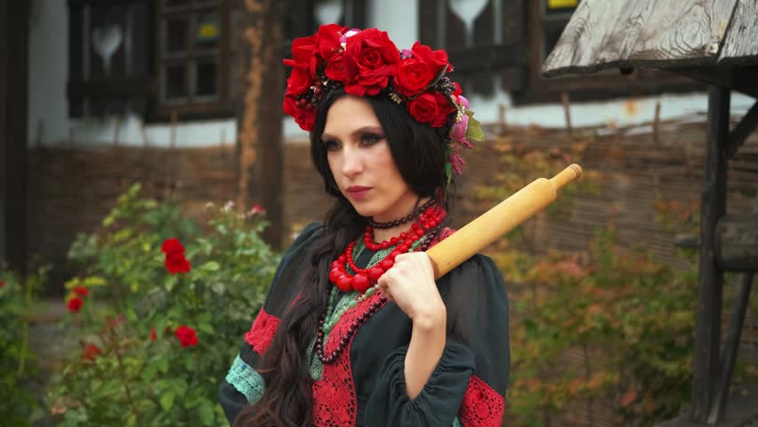 Fantasy portrait warlike evil woman with bat, holding in hands threatening looks at camera. black dress red beads wreath roses crown poppy flowers. angry wife threatens her husband with rolling pin.