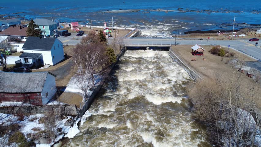 Dynamic aerial scene of fast-moving rapids rushing beneath a coastal bridge and community life in the village of St-Ulric-de-Matane along the St. Lawrence coastline. Quebec, Canada, 2026.