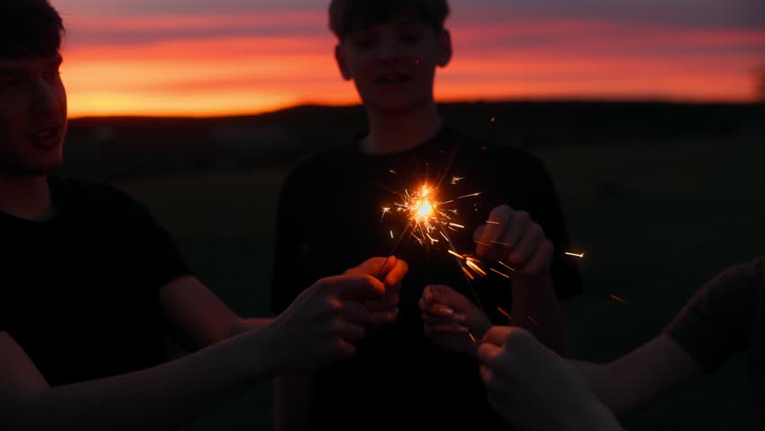 A group of young friends holding and pointing at a bright glowing sparkler firework with flying sparks against a deep red colorful twilight sky backdrop.