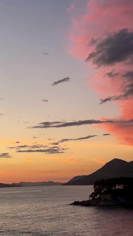 Vibrant sunset sky above Dubrovnik rocky coastline with calm Adriatic Sea, mountain silhouettes, and dramatic pink clouds during peaceful evening atmosphere in Croatia.