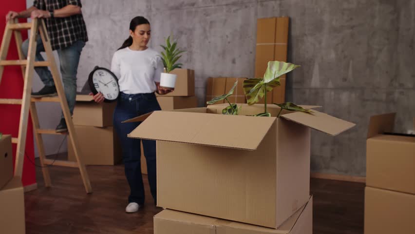Young woman carefully placing a clock and plants into a cardboard box while moving house
